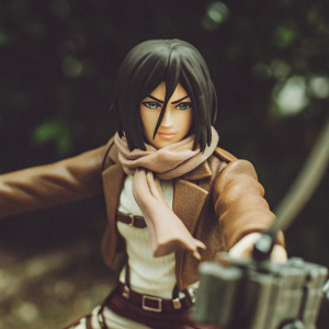 Mikasa Akerman, a detailed action figure. Green eyes, dark hair styled in a bob cut. Standing in a fierce pose. Wearing a brown jacket and a scarf.