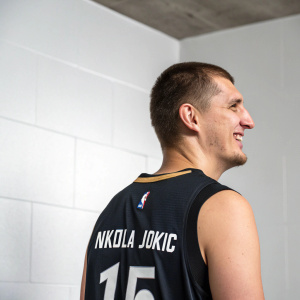 Nikola Jokic, with a smile, standing alone in a minimalist studio setting. His jersey is black with white text, highlighting his name and number.