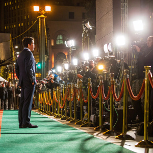 Rami Malek on a vibrant green carpet, wearing a sharp navy suit. Surrounded by flashing cameras and excited fans during a glamorous Hollywood awards night.