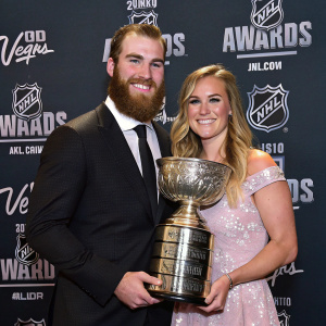 Ryan O'Reilly and Dayna Douros, smiling and holding a glittering trophy. The dark backdrop features shiny NHL Awards logos.