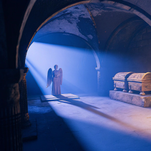 A 3D art piece from 'Rise of the Tomb Raider'. A crypt scene with an angel statue, ancient tomb, and arches. Dim lighting with rays of light casting shadows. Stone floor and walls. Tomb in the forefront.