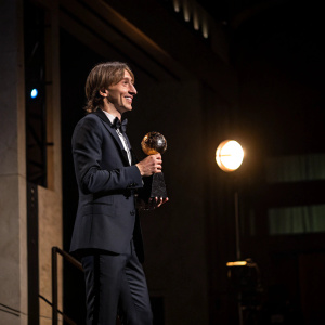 Luka Modric, in a formal suit, receiving award. Dimly lit room, spotlight on him.