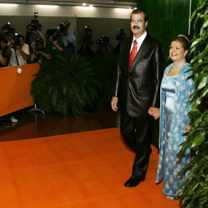 Former Mexican President Vicente Fox and his wife Marta Sahagun de Fox at a grand charity gala. Vicente Fox is in a sleek dark suit with a vibrant red tie, while Marta is elegantly dressed in a blue patterned gown. The couple is standing on a bustling red carpet.
