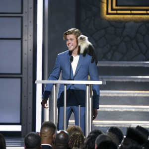 Justin Bieber at a roast event. Wearing a blue suit. Primate on shoulder. Crowded stage with blue backdrop and podium.