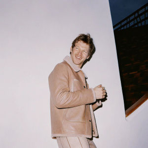 Julian Nagelsmann, a jovial expression, in a cozy brown leather jacket, standing against a blurred stadium backdrop.