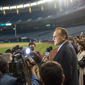 Joe Torre speaking at a stadium press conference. Crowd of reporters and camera flashes.