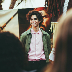 Cameron Boyce, smiling, with curly hair and wearing a pink shirt and green jacket, poses in front of a 'Pirates of the Caribbean: Dead Men Tell No Tales' promotional backdrop at a press event in Los Angeles.