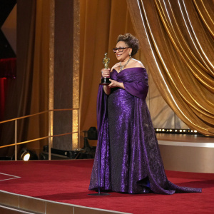 Ruth E Carter accepting award. She is wearing a beautiful purple dress. Standing on the red carpet at the Oscars. Behind her, the Dolby Theatre's golden curtains are displayed