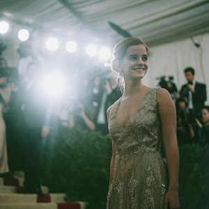Emma Watson at a glamorous gala event. Wears a stunning gold dress with intricate beading. Surrounded by bright lights and paparazzi.