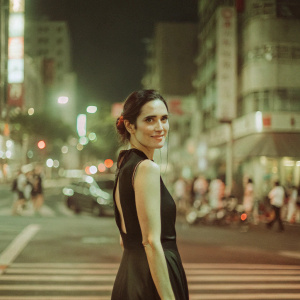 Jennifer Connelly, starring at the camera with a soft smile. Wearing a sleek black dress. Dark hair styled in a low bun. Against a backdrop of bustling city lights at night.