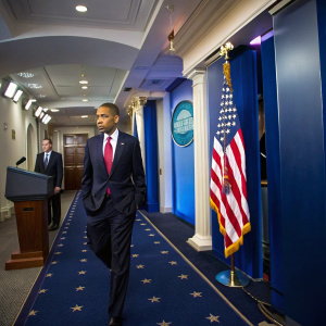 Barack Obama, in a navy suit and red tie, walking down a grand hallway. Blue walls adorned with American flags. Jay Carney stands nearby, both are at a White House press conference.