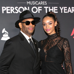 Jimmy Jam and Bella Harris at the MusiCares Person of the Year gala. Jimmy in a classic suit, sunglasses, and black fedora. Bella in a sleek, form-fitting black lace dress. Black backdrop with event logos and text.