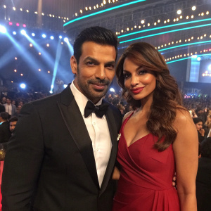 John Abraham and Bipasha Basu at the filmfare awards. John in a black tuxedo, Bipasha in a deep red gown. Flashing lights and audience in background.