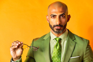 Middle Eastern man with a shaved head and a neatly trimmed goatee, wearing a suit and tie, holding a pair of eyeglasses in his hand