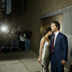 Formal event snapshot featuring Matthew McConaughey in a navy blue suit with a white dress shirt and burgundy tie. His wife Camila Alves McConaughey in a shimmering silver gown. They stand on a bustling sidewalk with paparazzi and fans