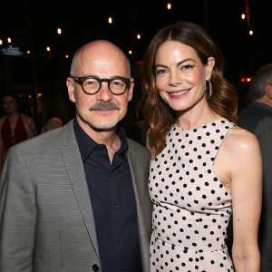 Clark Middleton and Michelle Monaghan at the awards night. Clark with a bald head, glasses, mustache in a dark shirt and gray blazer. Michelle with a big smile in a polka-dot dress. Dimly lit background with people around