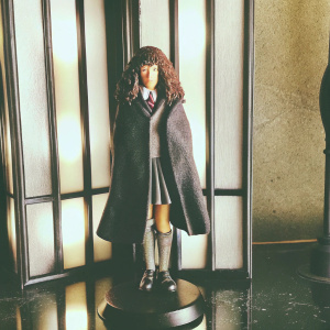 High-resolution render of Hermione Granger figurine. Dark cloak, curly dark hair, standing on a black base. The background is a minimalist design with sleek black and white accents.