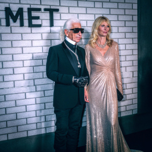 Karl Lagerfeld and Claudia Schiffer at the Met Gala. Lagerfeld in black suit and sunglasses. Schiffer in shimmering gold gown. White brick wall backdrop with 'Met' logo