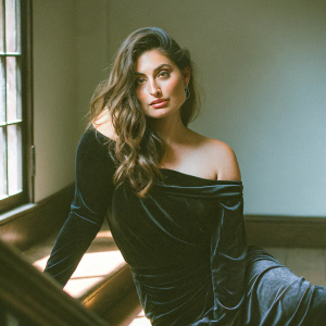 Caucasian actress Gabriella Wright, medium complexion, long wavy brown hair. Wears black velvet off-the-shoulder dress. Confident pose in a professional studio setting with soft lighting casting subtle shadows on plain light-colored background. Close-up portrait emphasizing upper body and attire.