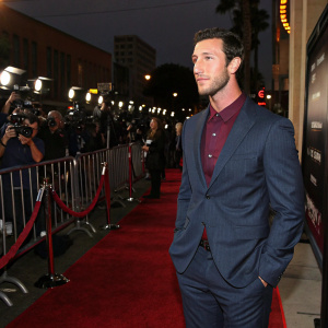 Pablo Schreiber, dressed in a dark blue suit with maroon shirt, posing alone at a Hollywood premiere. Red carpet with flashing cameras in the background.