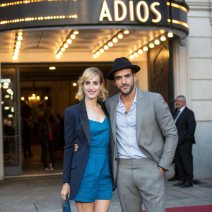 Natalia de Molina and Mario Casas attending the Madrid premiere of 'Adiós'. Natalia in a blue dress and blazer, Mario in a light shirt, black hat and grey suit. They pose in front of the movie theater marquee.